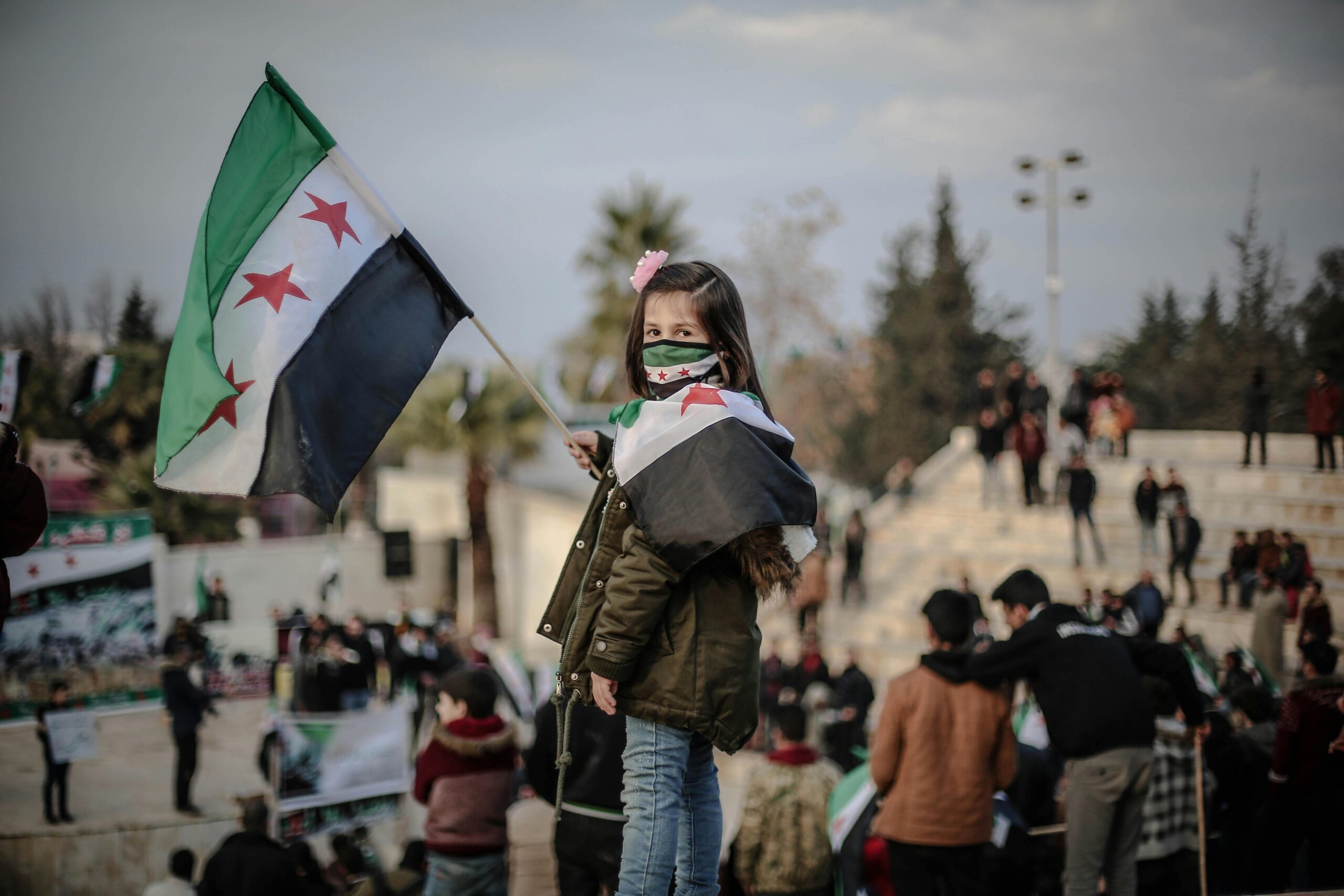 A young girl in Idlib, Syria, holds a flag during a protest rally, highlighting social issues.