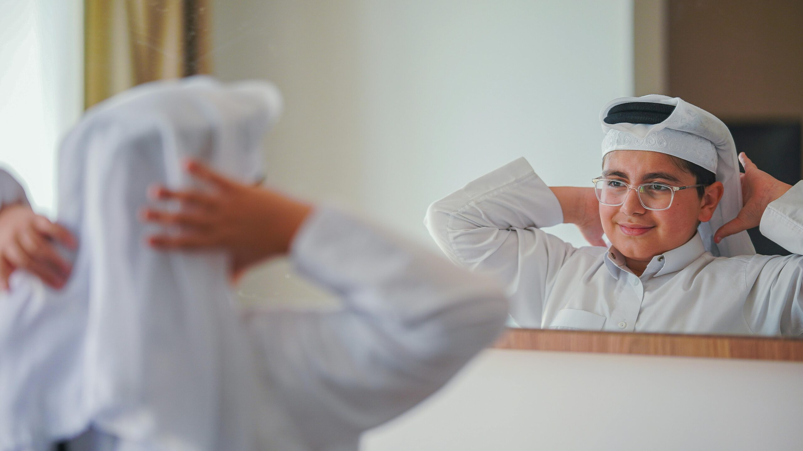 Arab boy wearing traditional attire looks in mirror, fixing headwear indoors.