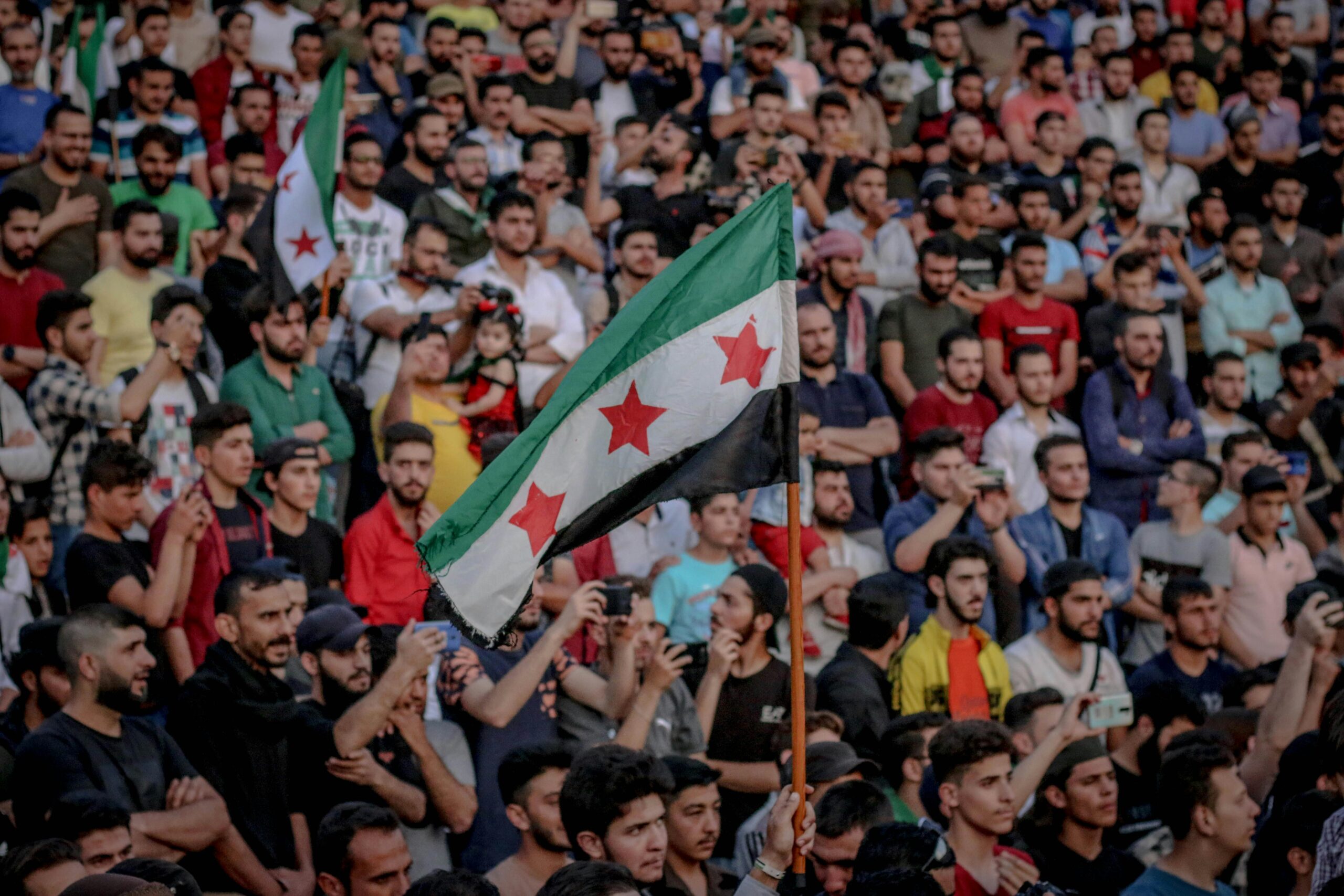 A large crowd gathers in protest, waving Syrian flags with red stars and expressing unity.